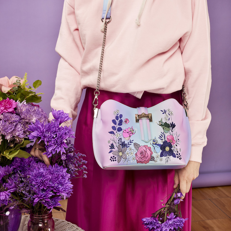 Image of someone wearing a pink sweater and skirt standing next to a bouquet of flowers and wearing the Loungefly Disney Sleeping Beauty 65th Anniversary Floral Ombre Crossbody Bag 